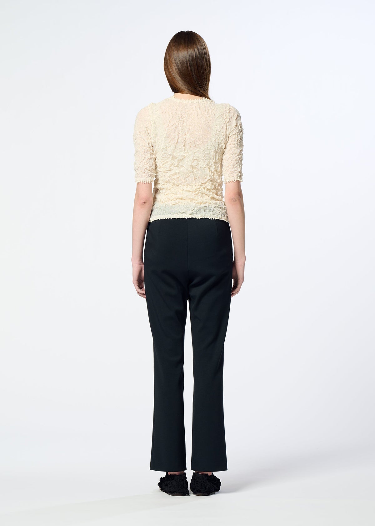 A person with long brown hair faces away from the camera, wearing HaaT's PONTE JERSEY textured short-sleeve cream top with flowy pants and black shoes, embodying relaxed comfort against a plain white background. view 8 of 8