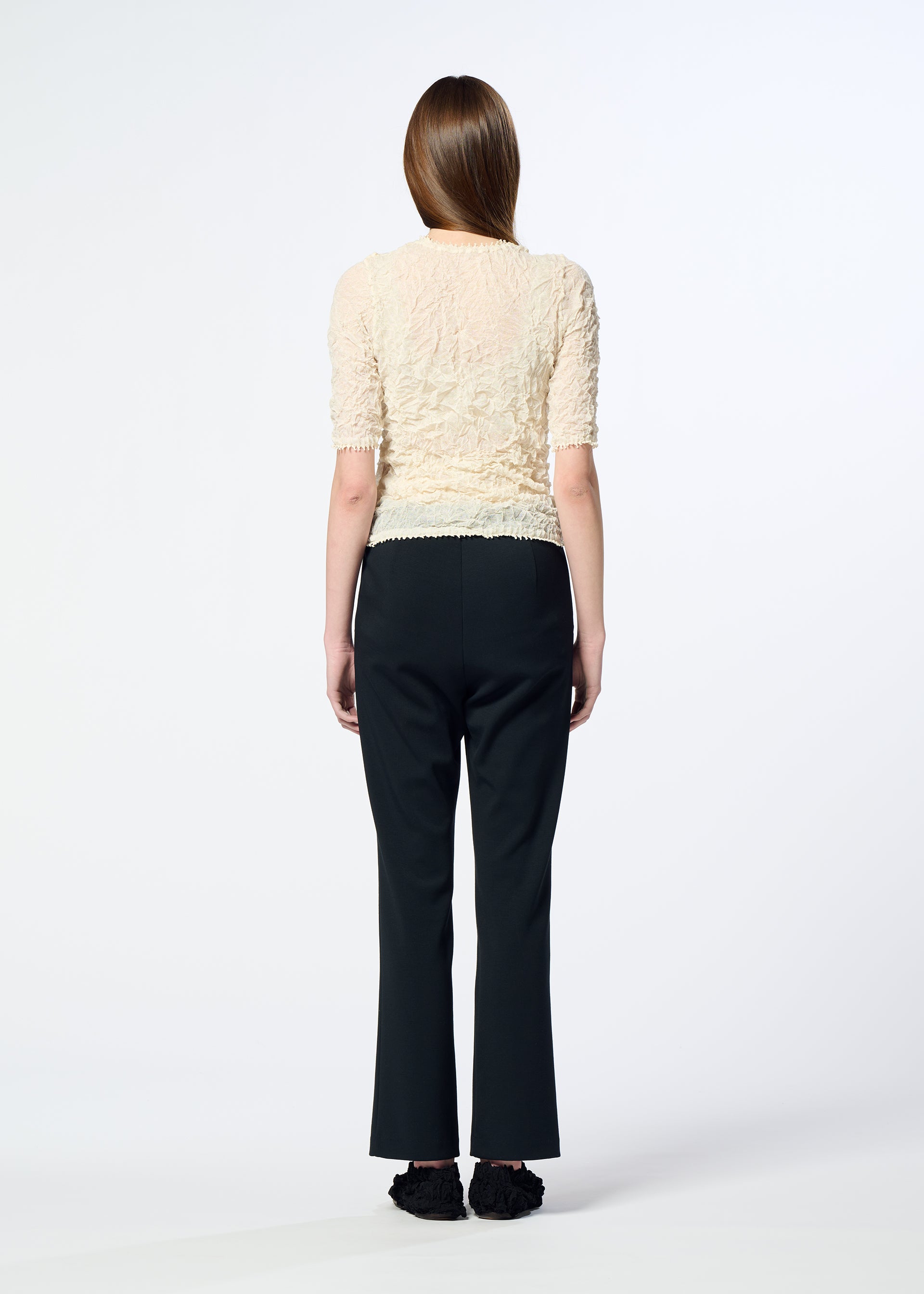 Zoomed view A person with long brown hair faces away from the camera, wearing HaaT's PONTE JERSEY textured short-sleeve cream top with flowy pants and black shoes, embodying relaxed comfort against a plain white background.