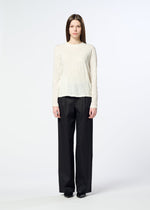 A person stands facing the camera, wearing the HaaT W/P WASHER—a cream round-neck long-sleeved top crafted from polyester wool jersey with a subtle wrinkled finish—paired with loose black pants and black shoes, against a plain white background.