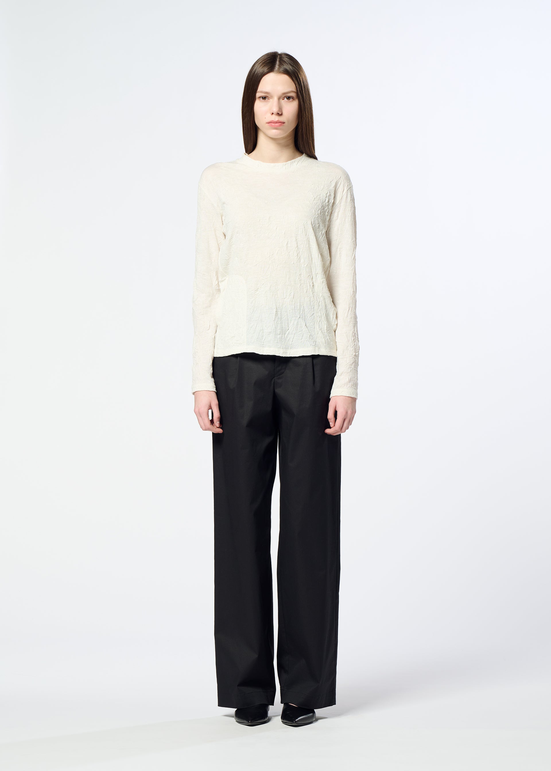 Zoomed view A person stands facing the camera, wearing the HaaT W/P WASHER—a cream round-neck long-sleeved top crafted from polyester wool jersey with a subtle wrinkled finish—paired with loose black pants and black shoes, against a plain white background.