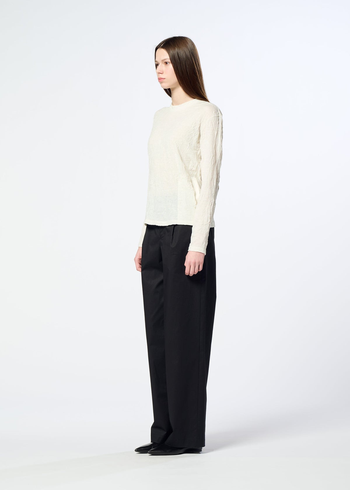 A woman with straight brown hair is seen in profile against a plain white background, wearing HaaT’s W/P WASHER—a round-neck, long-sleeved top crafted from polyester wool jersey with a subtle wrinkled finish—paired with loose black trousers. view 7 of 8