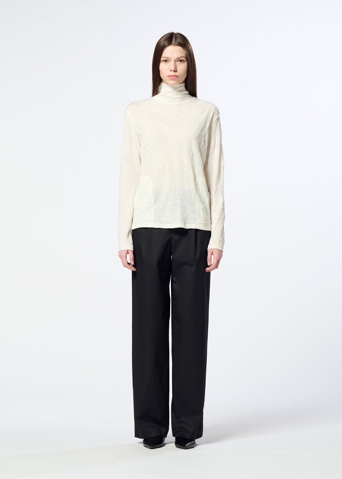 A person stands facing forward against a plain white background, wearing HaaT’s W/P WASHER cream wool jersey turtleneck sweater and loose black trousers. view 6 of 8