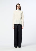 A person stands facing forward against a plain white background, wearing HaaT’s W/P WASHER cream wool jersey turtleneck sweater and loose black trousers.