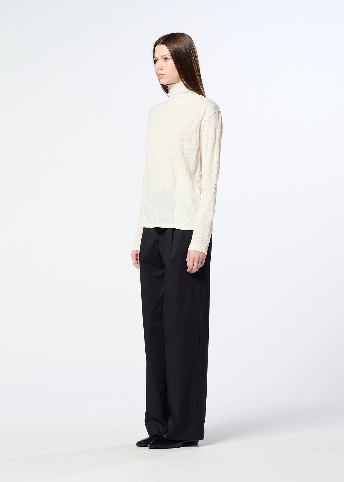 A person stands sideways against a plain white background, wearing the HaaT W/P WASHER cream wool jersey turtleneck sweater, loose black trousers, and black shoes. view 7 of 8
