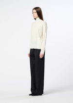 A person stands sideways against a plain white background, wearing the HaaT W/P WASHER cream wool jersey turtleneck sweater, loose black trousers, and black shoes.