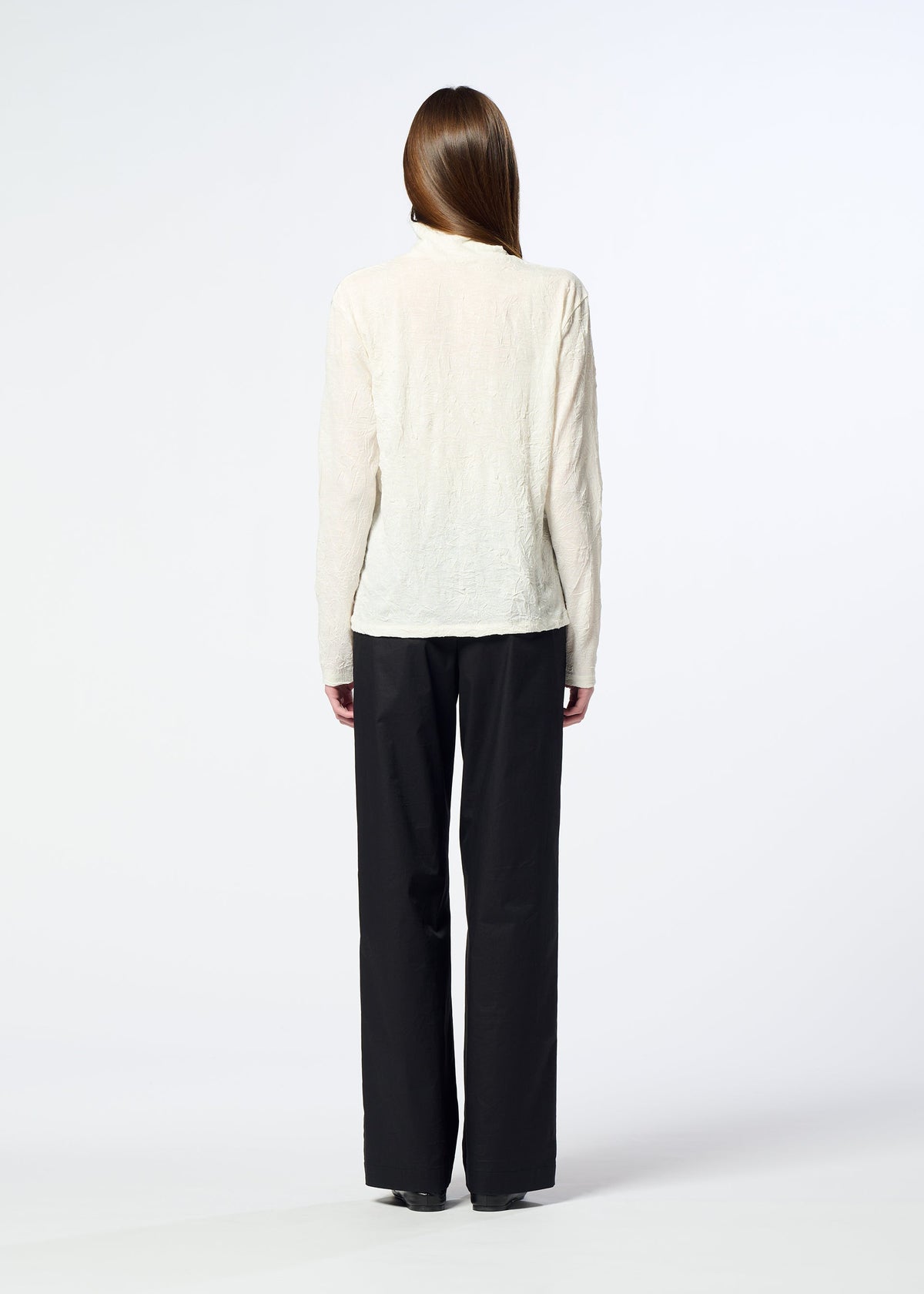 A person with long hair, seen from behind, is wearing HaaT’s W/P WASHER cream-colored high-neck wool jersey top and loose black trousers, standing against a plain white background. view 8 of 8
