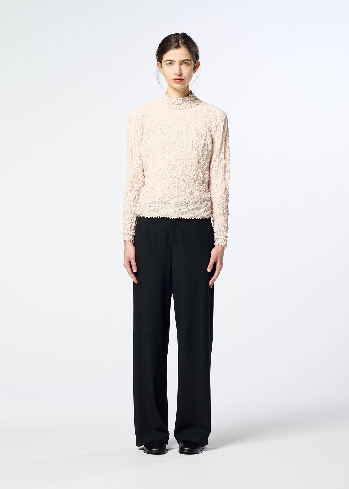 Zoomed view A person stands facing forward wearing the KUMO SHIBORI by HaaT, a textured light pink tricot jersey high-neck top, with loose black trousers and black shoes against a plain, light background.