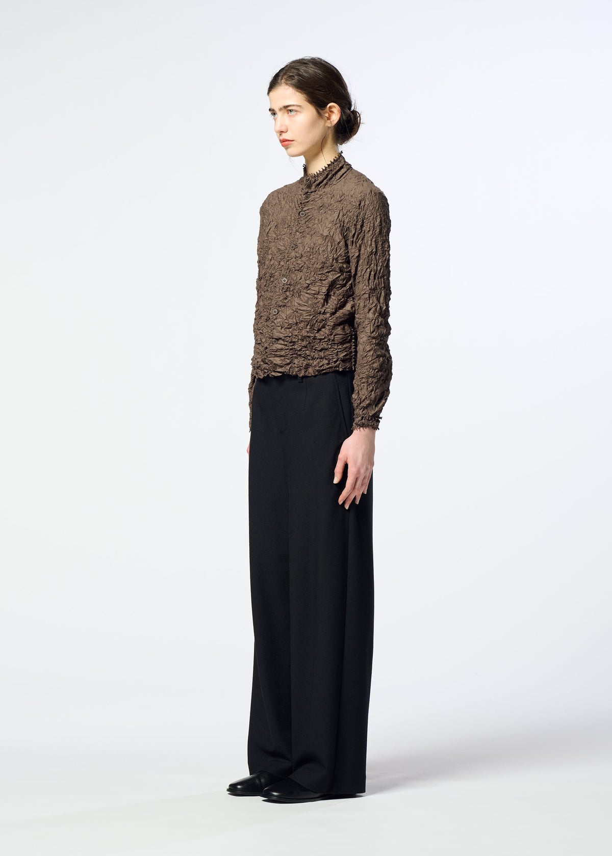 Zoomed view A person stands in profile against a plain light background, wearing the HaaT KUMO SHIBORI textured brown high-neck cardigan with wide-leg black pants and black shoes.