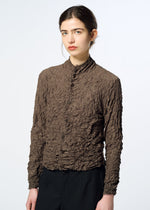 A person with long dark hair in a low bun wears the HaaT KUMO SHIBORI textured, brown crinkled long-sleeve shirt with black pants against a plain light background.
