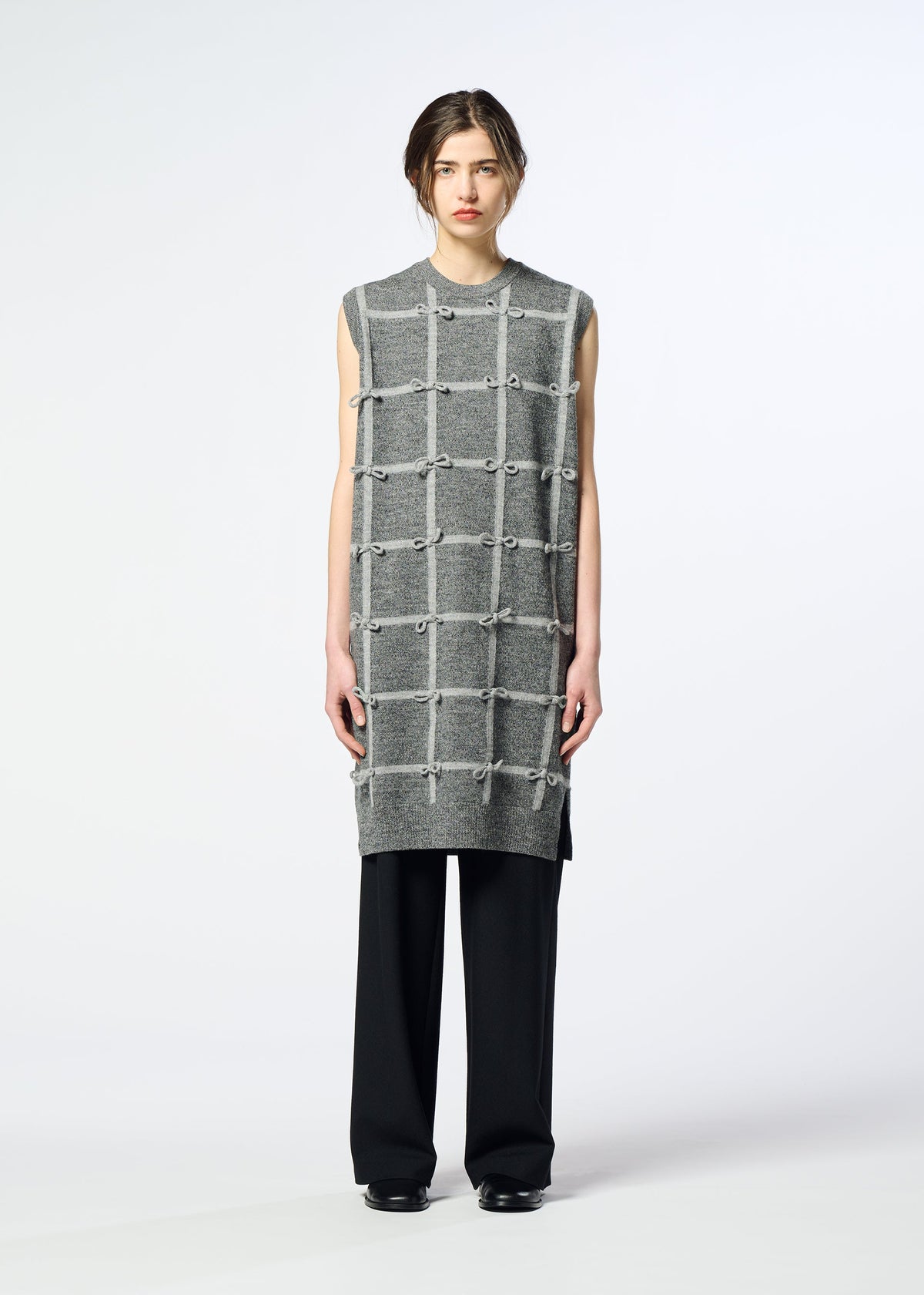 A person stands facing forward, wearing the HaaT RIBBON KNIT—a sleeveless gray tunic with a grid pattern and tied ribbons—over black wide-leg pants and black shoes, against a plain light background. view 5 of 8