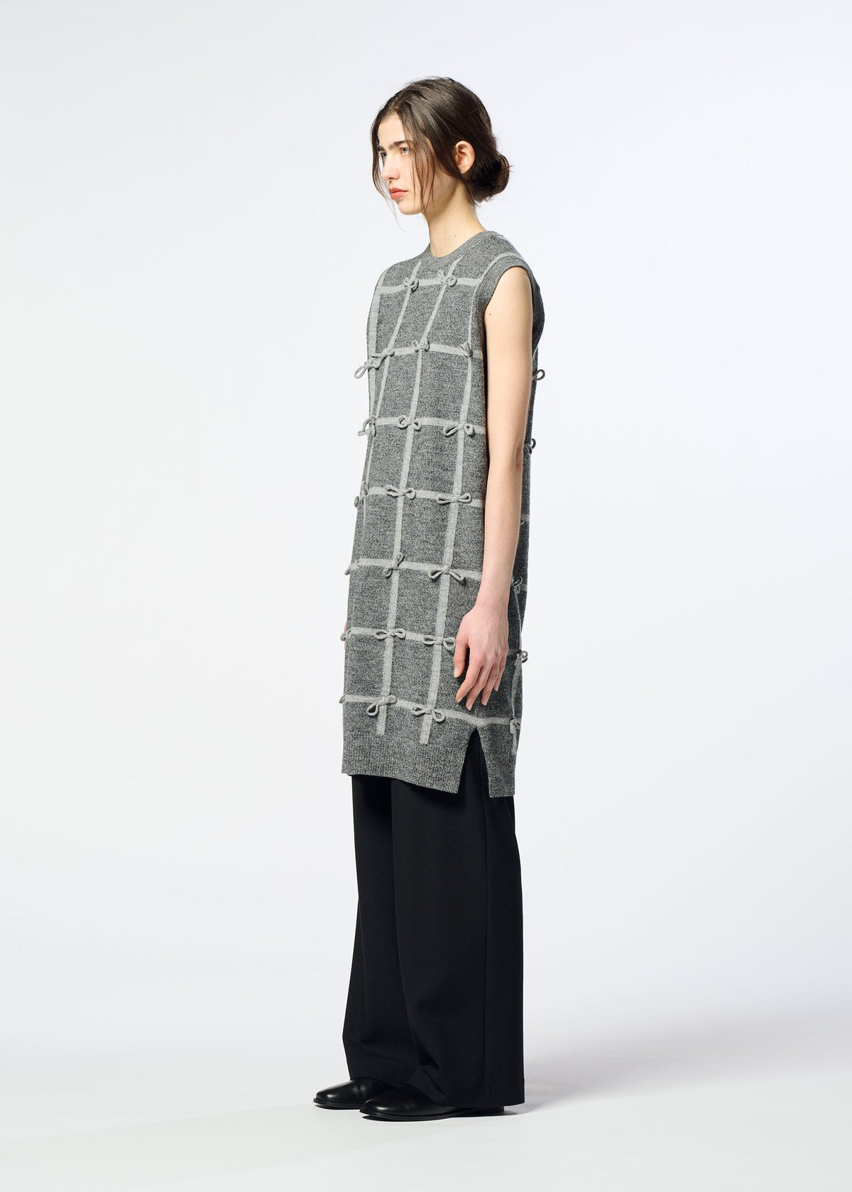 Zoomed view A woman stands in profile wearing the HaaT RIBBON KNIT, a long, sleeveless grid-patterned wool dress with knots, over wide black pants and shoes against a plain light background.