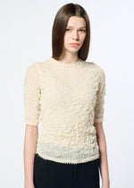 A young woman with straight brown hair wears the HaaT KUMO SHIBORI KNIT SHEER, a light beige ramie-blend short-sleeve top, paired with black pants against a plain white background.