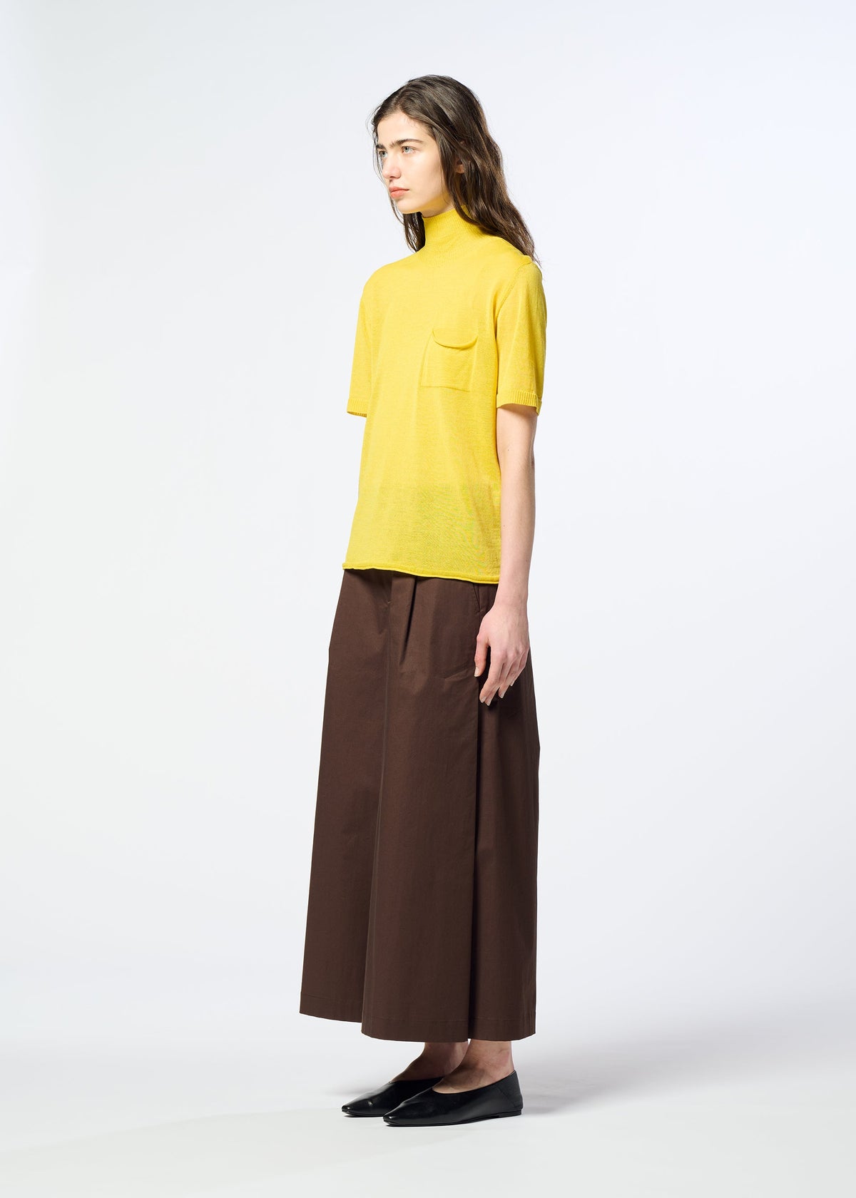 A person stands sideways against a plain white background, wearing HaaT’s AIRY POCKET LAYERS—a yellow short-sleeved knit turtleneck top with a pocket—paired with brown wide-leg pants and black flats. The minimalist knit construction of the AIRY POCKET LAYERS suggests it could be made from a soft polyester wool blend, providing both comfort and style. view 7 of 8