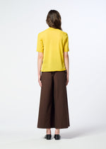 A person stands facing away, dressed in HaaT’s AIRY POCKET LAYERS—a short-sleeved knit yellow shirt—paired with wide-legged brown pants made from a polyester wool blend and black shoes, set against a plain, light background.
