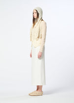 A woman stands in profile against a plain background, wearing HaaT’s KUMO SHIBORI KNIT SHEER, a textured cream-colored ramie-blend hooded top, paired with a long white skirt and matching slip-on shoes.