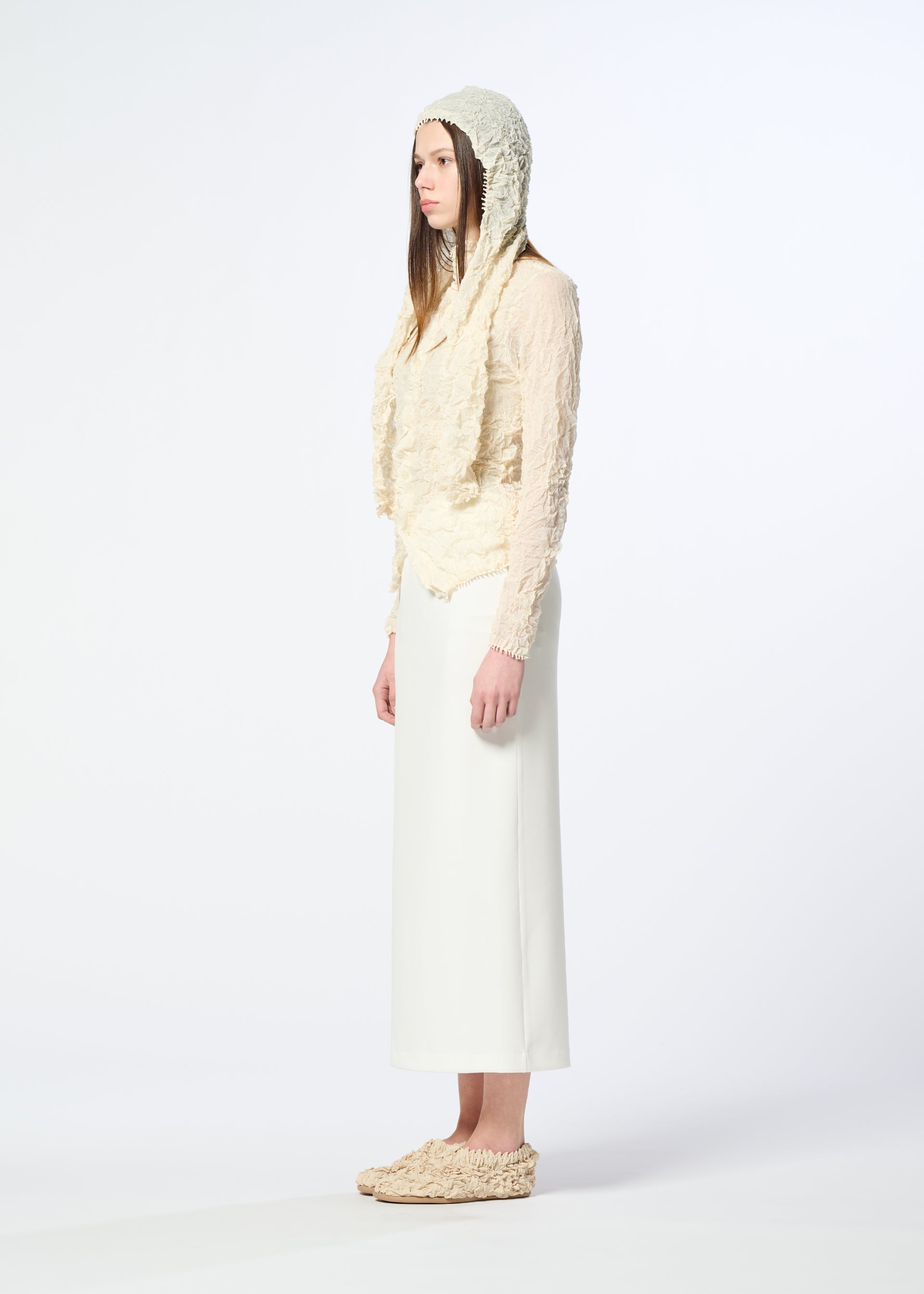 Zoomed view A woman stands in profile against a plain background, wearing HaaT’s KUMO SHIBORI KNIT SHEER, a textured cream-colored ramie-blend hooded top, paired with a long white skirt and matching slip-on shoes.