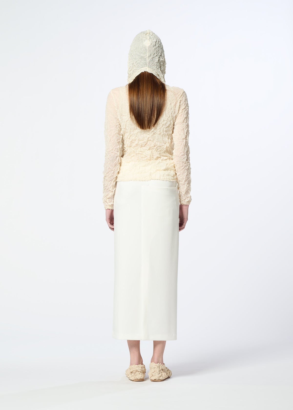 A person with long straight hair, facing away, wears the HaaT KUMO SHIBORI KNIT SHEER in textured cream ramie-blend with a hood, paired with a white ankle-length skirt and fluffy cream slippers against a plain white background. view 8 of 9
