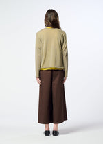 A person stands facing away, dressed in HaaT’s AIRY POCKET LAYERS—a tan minimalist knit sweater—layered over a yellow shirt, paired with wide brown pants and black shoes, against a plain white background.