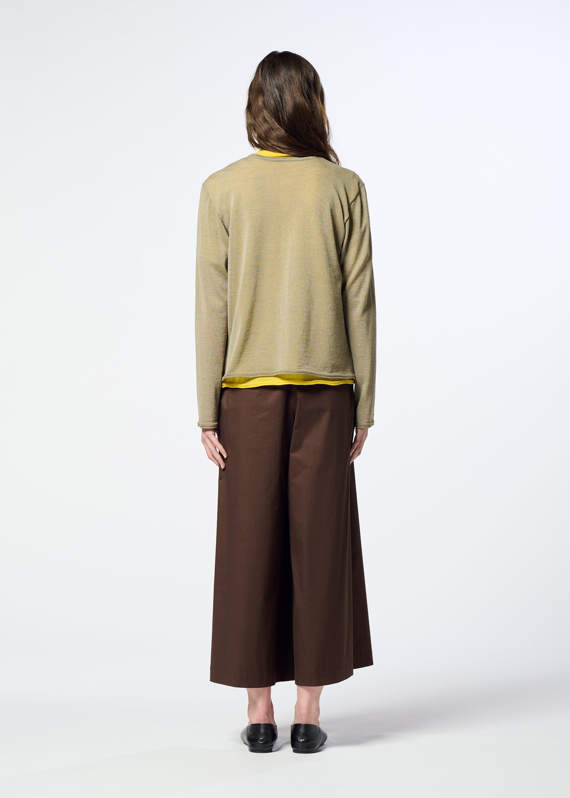 Zoomed view A person stands facing away, dressed in HaaT’s AIRY POCKET LAYERS—a tan minimalist knit sweater—layered over a yellow shirt, paired with wide brown pants and black shoes, against a plain white background.