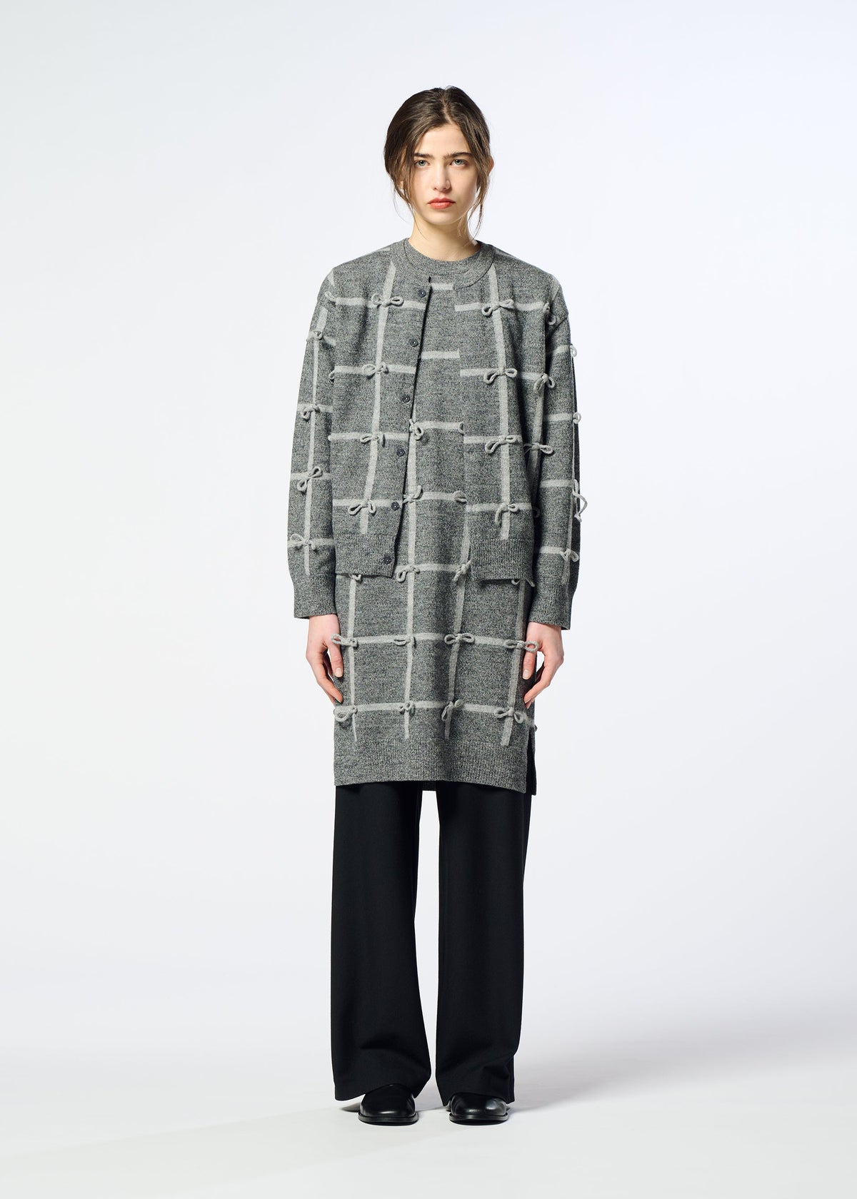 A person stands against a plain background wearing the HaaT RIBBON KNIT cardigan with knotted details, loose black pants, black shoes, and a neutral expression. view 5 of 7