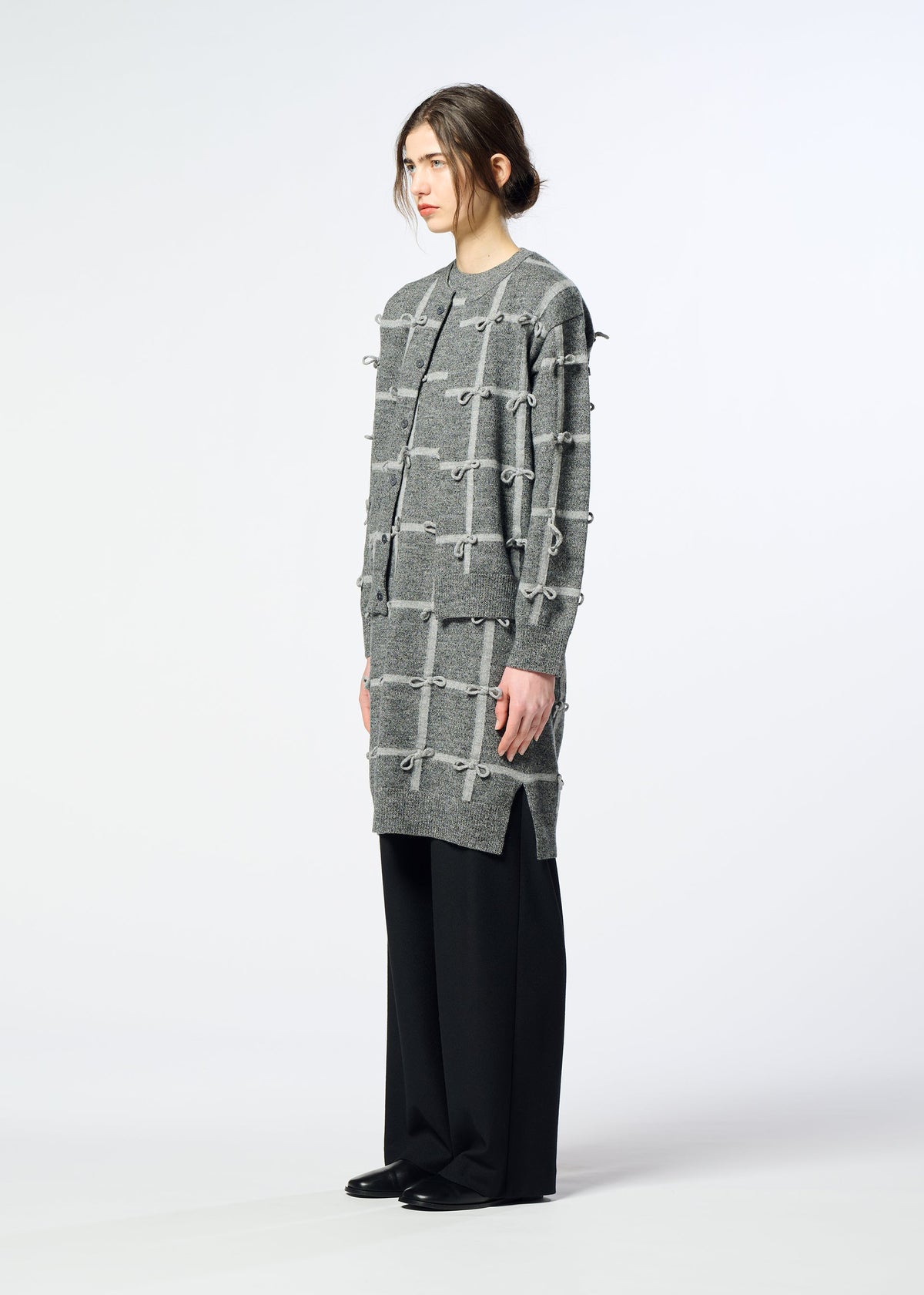 Zoomed view A person stands sideways in the HaaT RIBBON KNIT, a grey, grid-patterned sweater dress with knots, paired with black wide-leg pants and a long-sleeved knit cardigan against a plain white background.