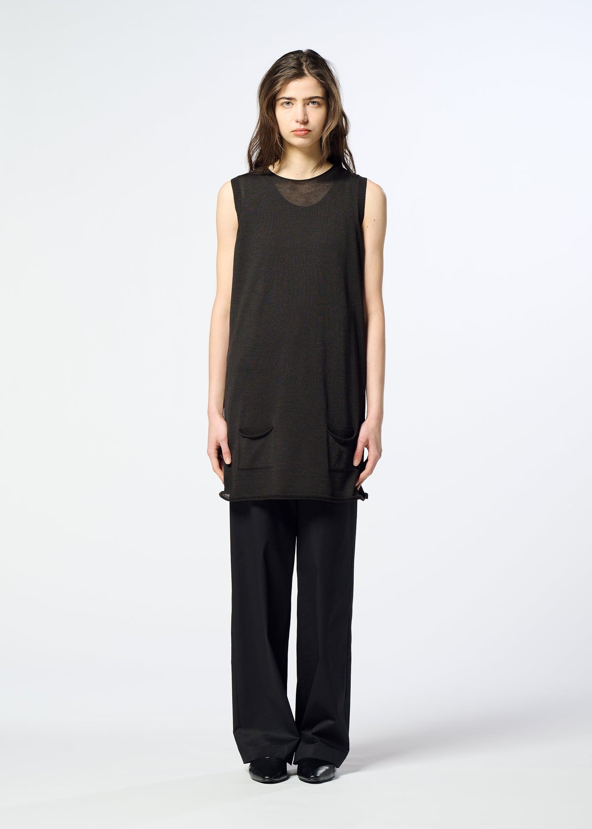 A woman stands facing forward, dressed in HaaT’s AIRY POCKET LAYERS—a sleeveless black tunic featuring two front pockets—paired with loose black pants for a chic layered look. The background is plain and white. view 6 of 8