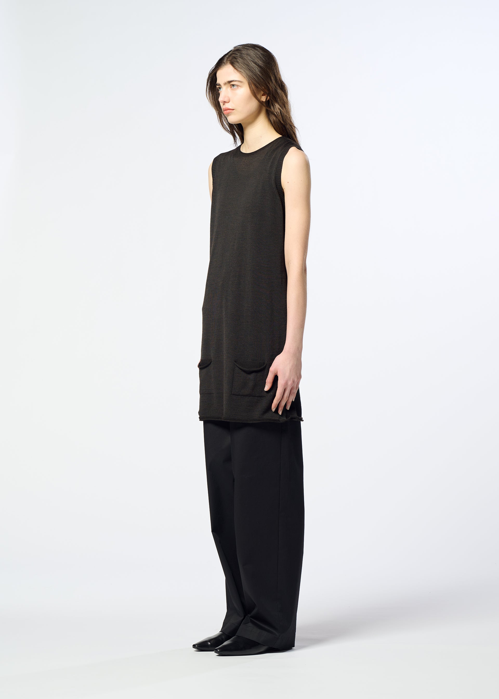Zoomed view A person with long brown hair stands sideways against a plain light background, wearing HaaT’s AIRY POCKET LAYERS—a minimalist black knit sleeveless tunic with front pockets—layered over wide-leg black pants and black shoes.
