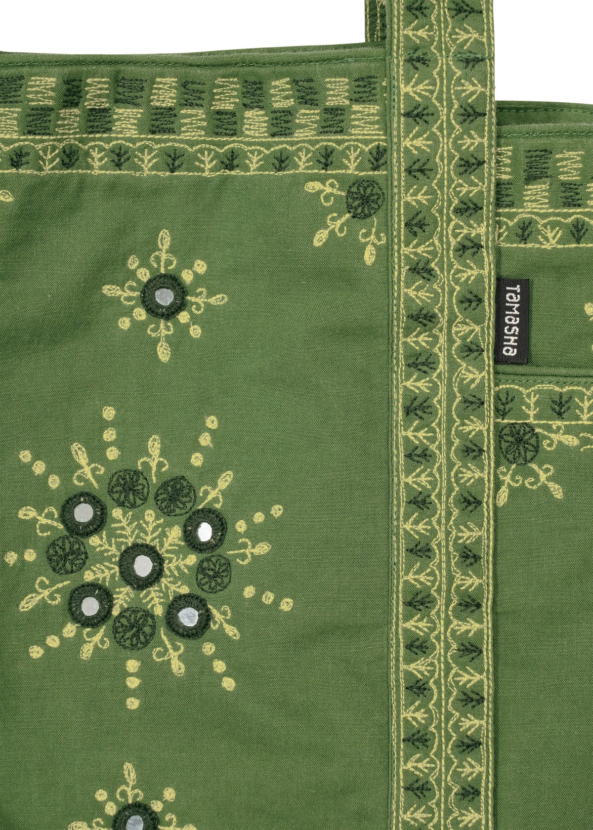 The DAZZLE MIRROR BOX by HaaT is a green fabric mini tote bag adorned with yellow and dark green Bhill embroidery patterns, mirror accents, and a decorative handle. Lovingly crafted by Indian artisans, it features a black label with white HaaT branding attached to the bag. view 7 of 8