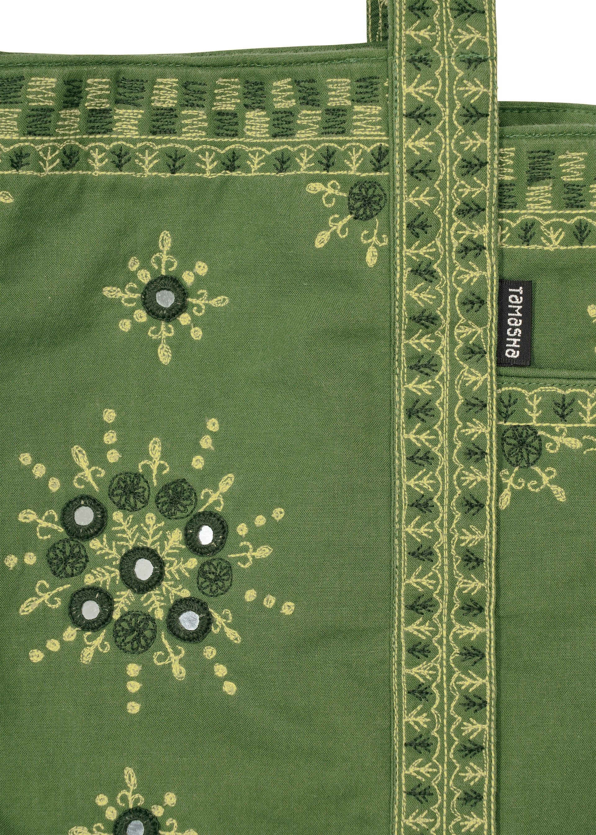 Zoomed view The DAZZLE MIRROR BOX by HaaT is a green fabric mini tote bag adorned with yellow and dark green Bhill embroidery patterns, mirror accents, and a decorative handle. Lovingly crafted by Indian artisans, it features a black label with white HaaT branding attached to the bag.