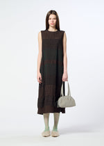 A woman stands against a white background, holding the BLOOM & PLAID BOSTON bag from HaaT—a light gray Boston bag. She wears a sleeveless, brown midi dress with subtle patterns, complemented by green tights and fluffy slippers. Her long, straight hair adds to the look, which is beautifully accented by intricate Bhil embroidery celebrating the craftsmanship of Indian artisans.