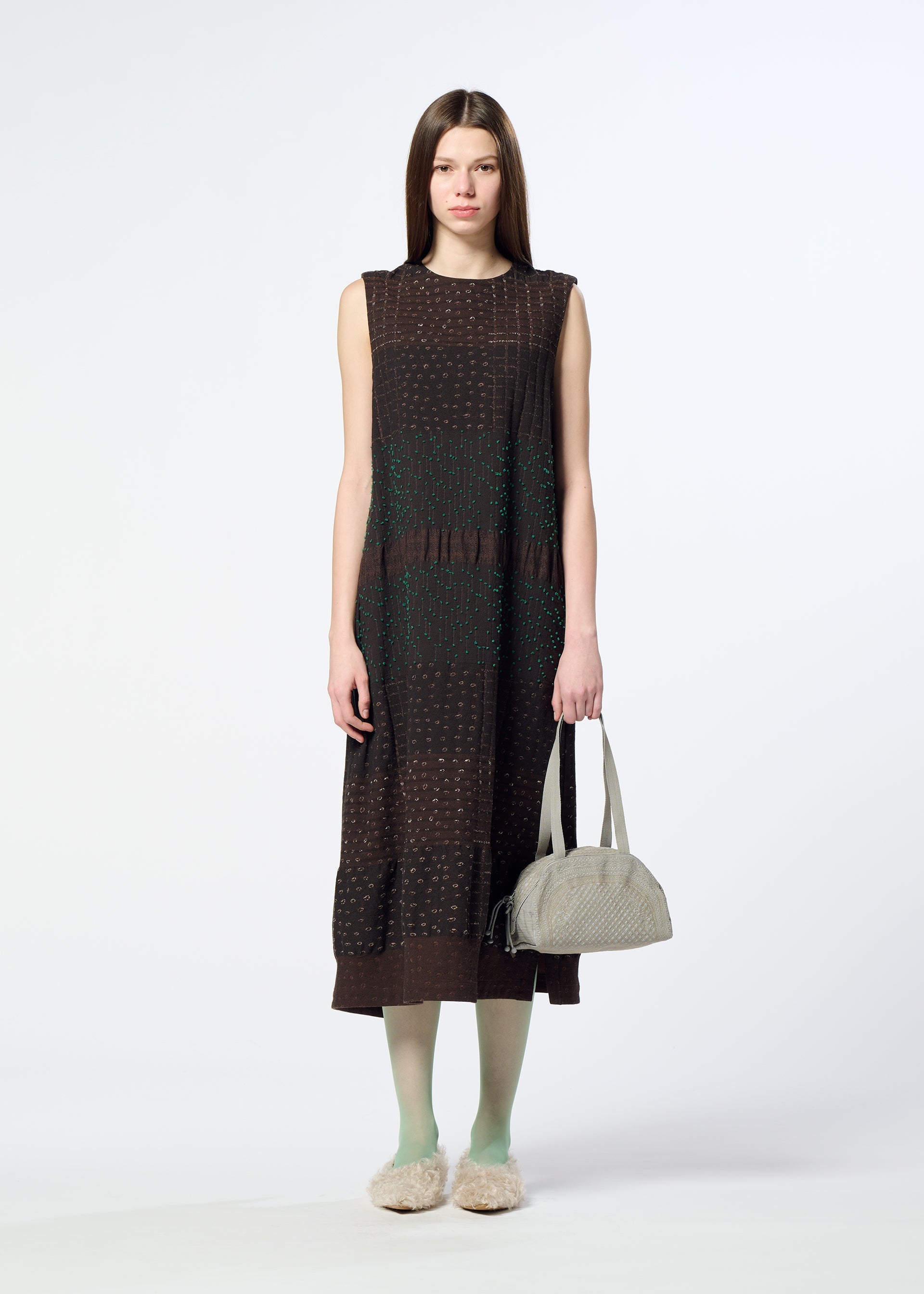 Zoomed view A woman stands against a white background, holding the BLOOM & PLAID BOSTON bag from HaaT—a light gray Boston bag. She wears a sleeveless, brown midi dress with subtle patterns, complemented by green tights and fluffy slippers. Her long, straight hair adds to the look, which is beautifully accented by intricate Bhil embroidery celebrating the craftsmanship of Indian artisans.