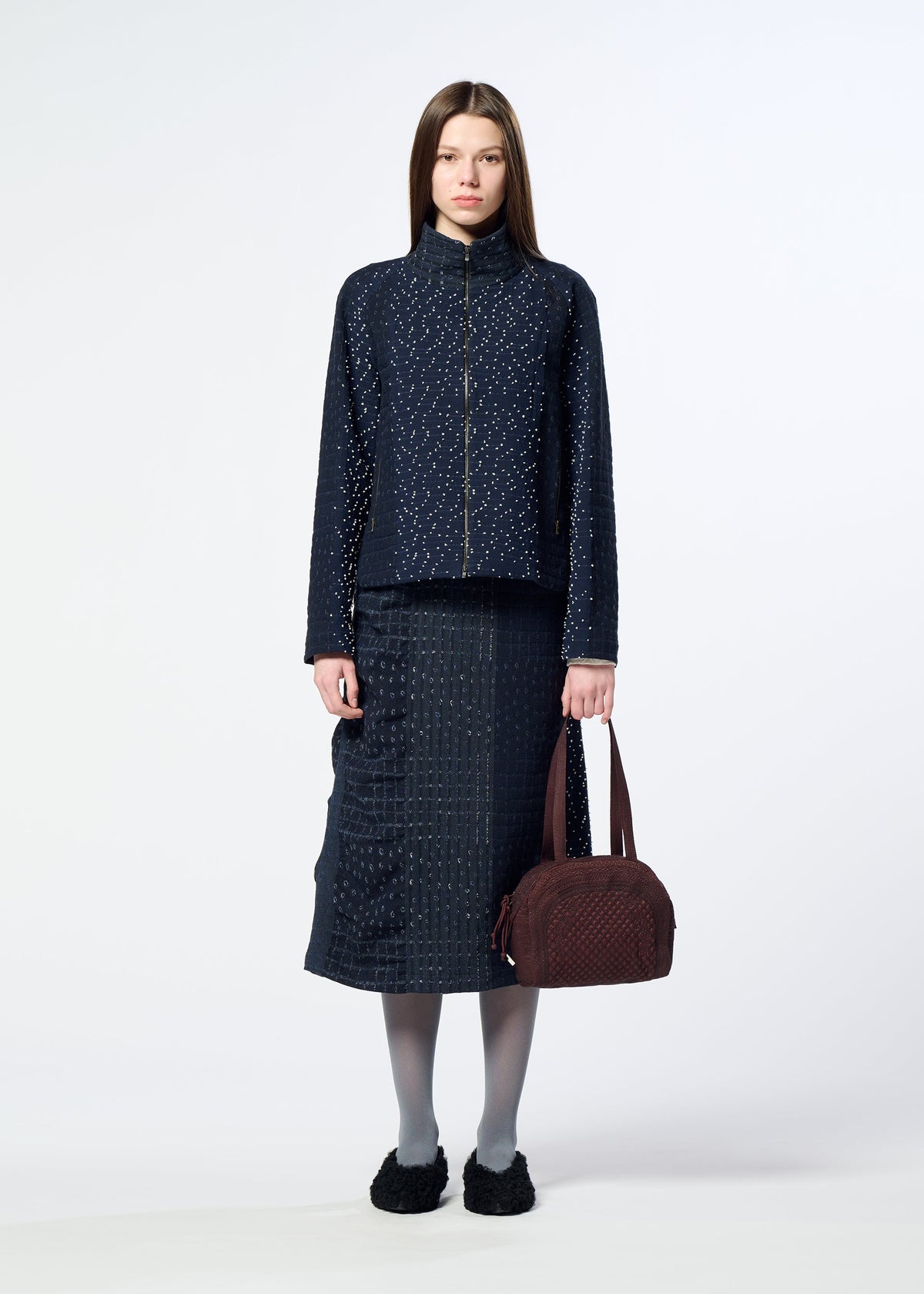 A woman stands facing forward against a plain white background, wearing a navy textured jacket and skirt, gray tights, and black shoes, while holding the HaaT BLOOM & PLAID BOSTON bag in maroon. view 7 of 7
