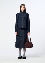 A woman stands facing forward against a plain white background, wearing a navy textured jacket and skirt, gray tights, and black shoes, while holding the HaaT BLOOM & PLAID BOSTON bag in maroon.