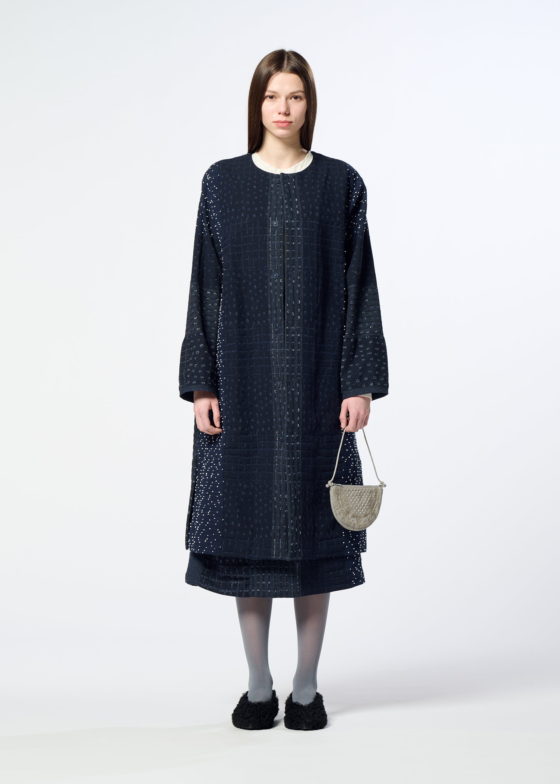 Zoomed view A woman stands against a plain background wearing a long, dark textured coat, matching skirt, gray tights, and black shoes, holding the BLOOM & PLAID BOSTON pouch by HaaT—crafted in India and adorned with intricate Bhill embroidery.