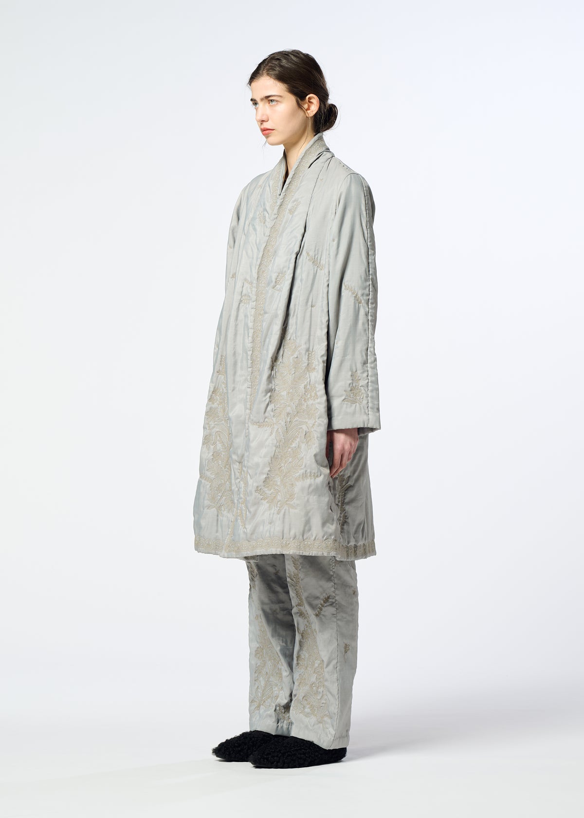 A person stands sideways wearing the DELICATE FLORAL BHILL by HaaT, a light gray oversized quilted coat with subtle botanical patterns and matching pants, paired with dark slippers against a plain white background. view 8 of 9