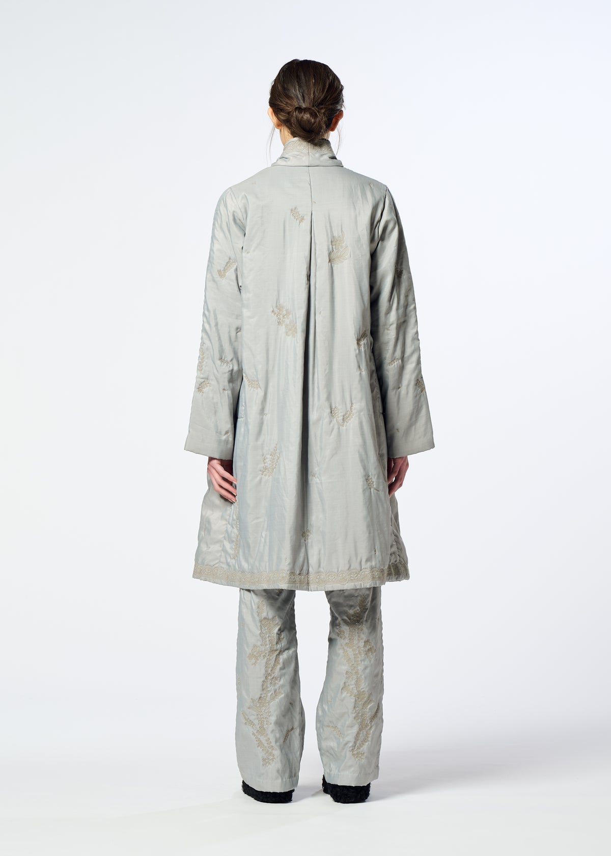 Zoomed view A person stands facing away, wearing HaaT’s DELICATE FLORAL BHILL light gray, knee-length evening coat and matching pants with subtle Bhill embroidery. Hair is tied back; background is plain white.