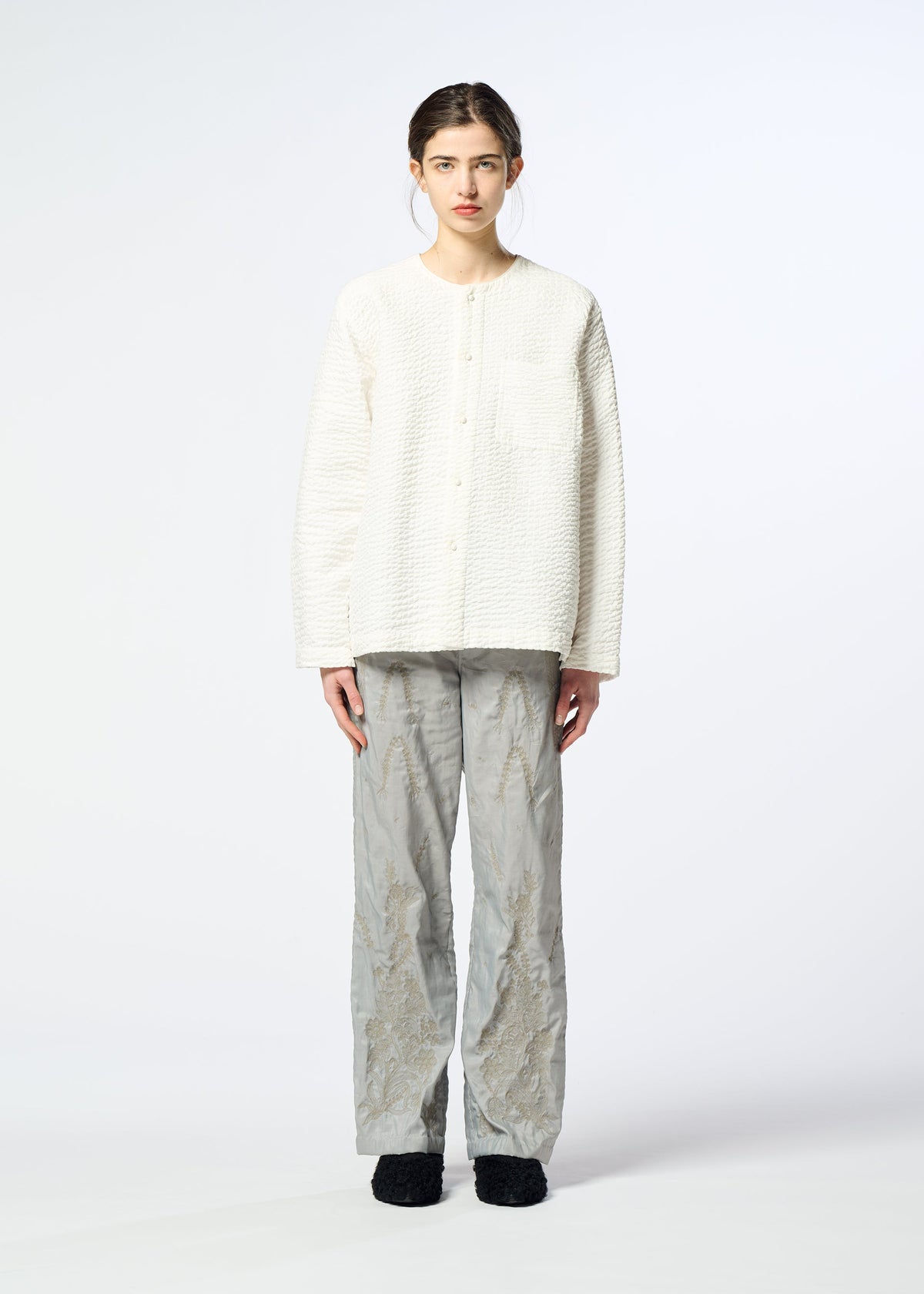 A person with straight brown hair tied back and a neutral expression wears the HaaT DELICATE FLORAL BHILL shirt, loose light gray pants, and black shoes against a plain white background. view 7 of 9