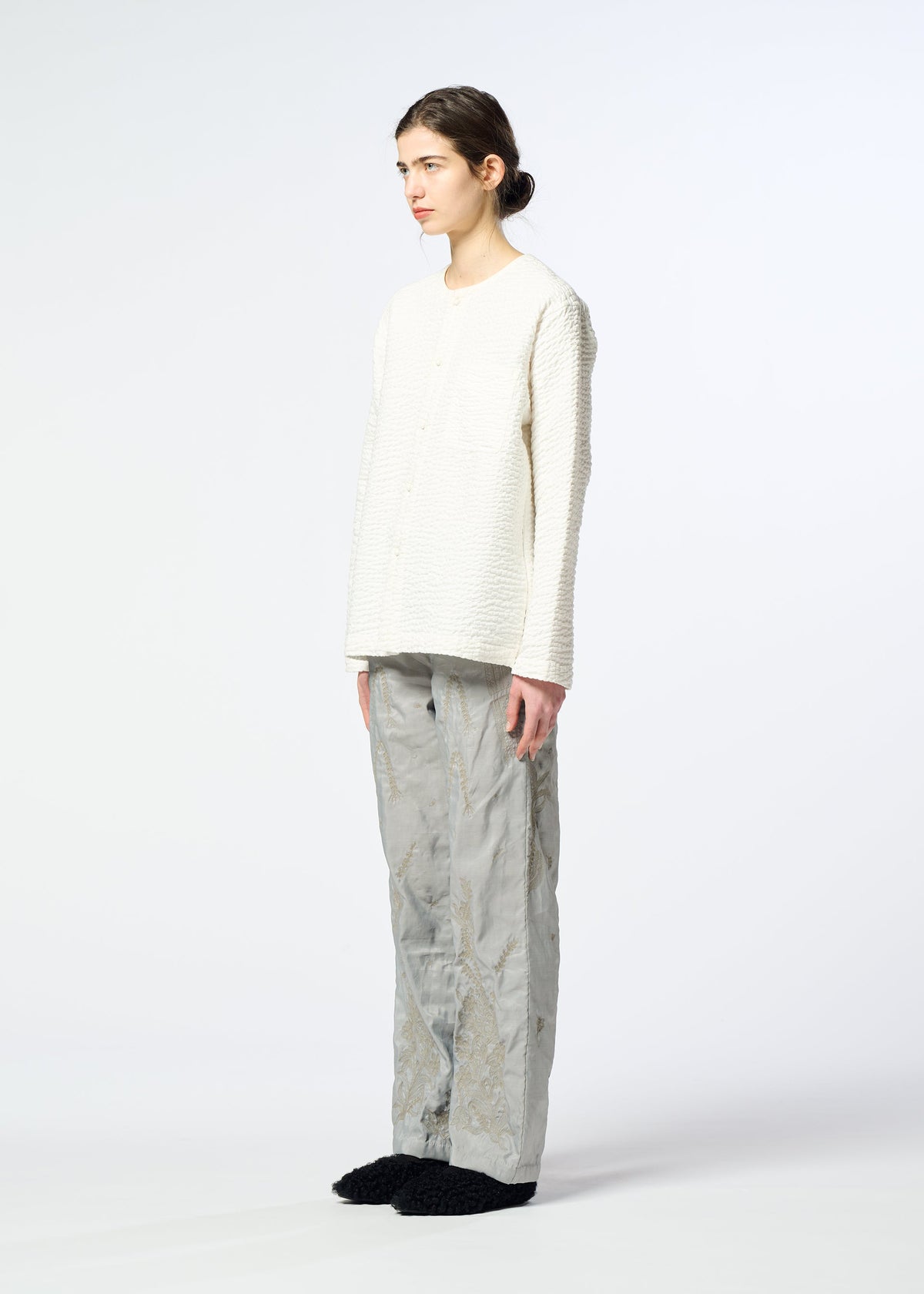 A person with long brown hair tied back stands sideways against a white background, wearing a textured white top, black slippers, and HaaT DELICATE FLORAL BHILL loose light gray pants with a faint botanical pattern. view 8 of 9