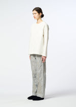 A person with long brown hair tied back stands sideways against a white background, wearing a textured white top, black slippers, and HaaT DELICATE FLORAL BHILL loose light gray pants with a faint botanical pattern.
