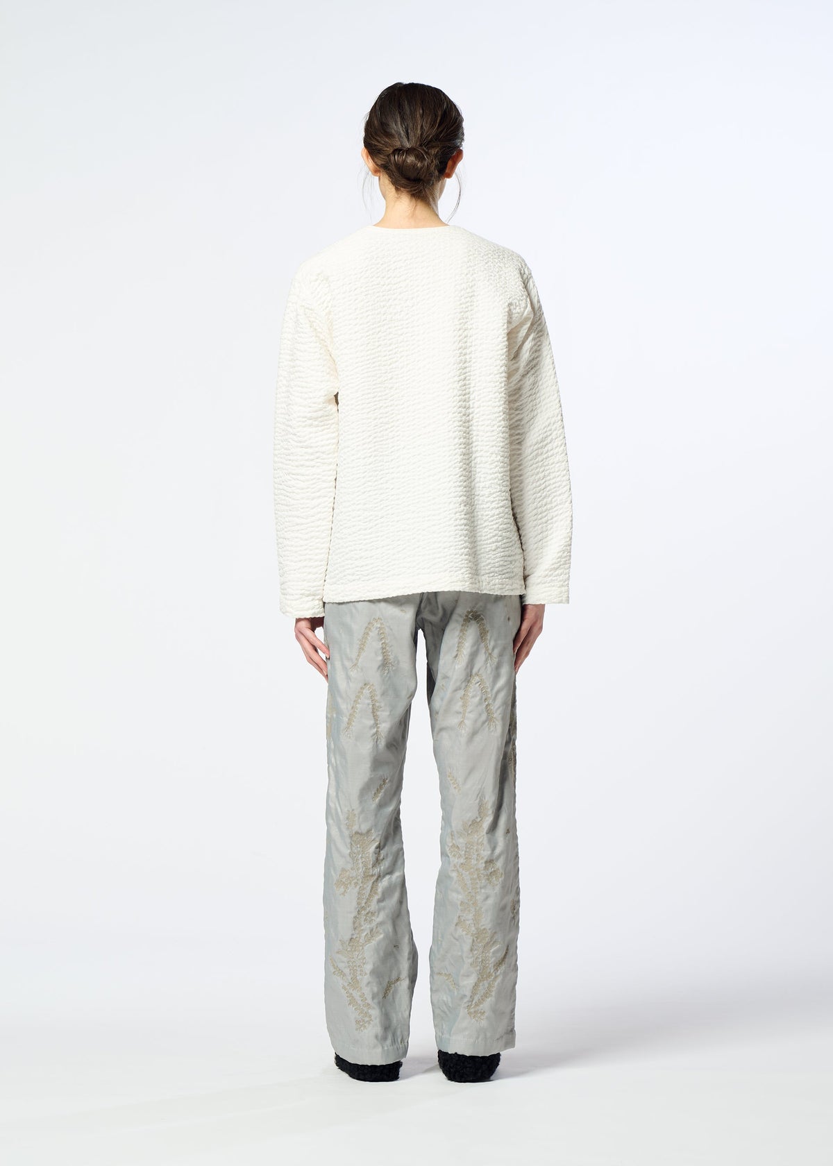 Zoomed view A person with dark hair in a low bun faces away, wearing a textured white sweater and HaaT DELICATE FLORAL BHILL loose, light gray nylon pants with a subtle botanical pattern against a plain background.
