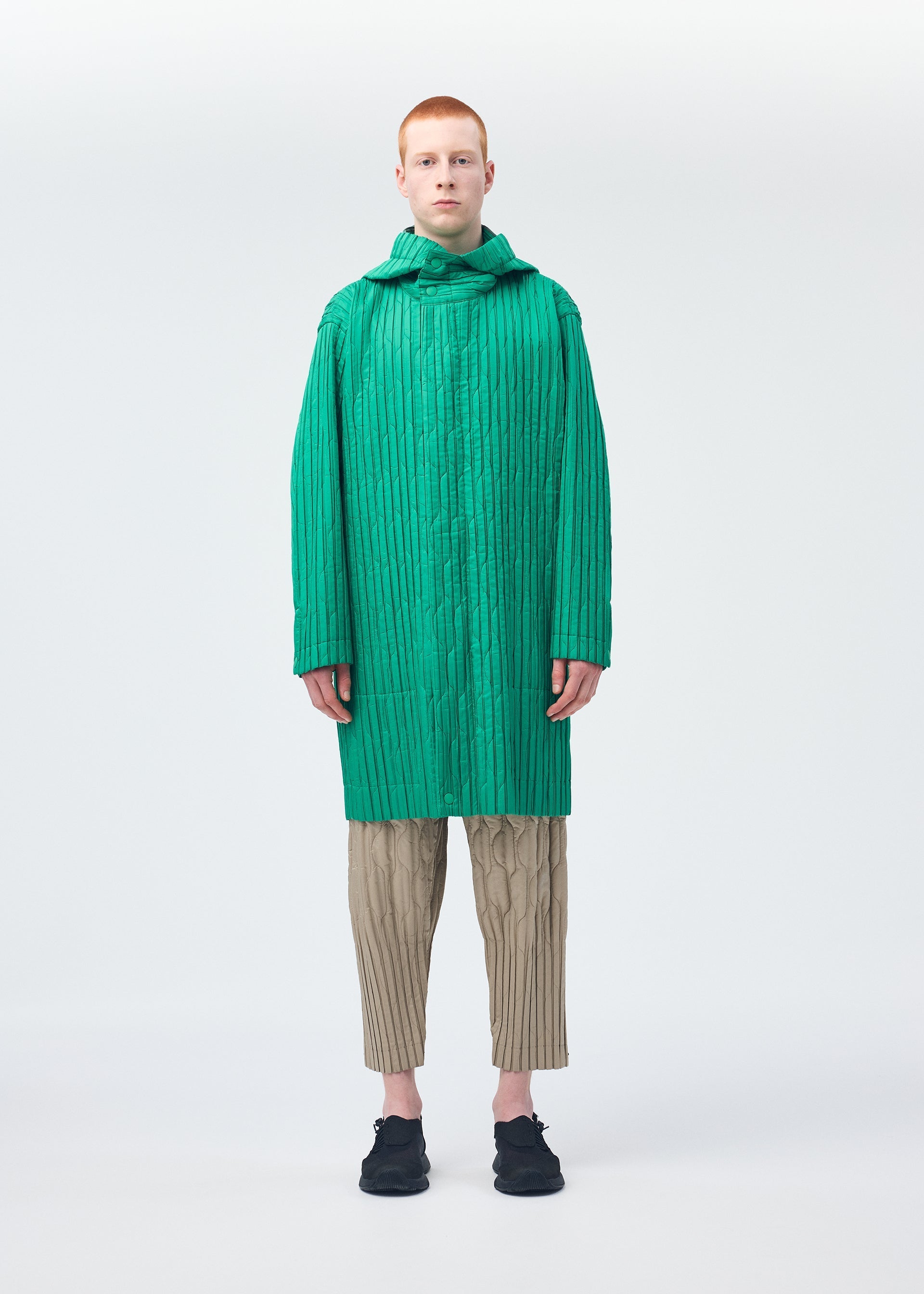 Zoomed view A person with short red hair models the HOMME PLISSÉ ISSEY MIYAKE PADDED PLEATS collection: a bright green textured long coat and matching scarf, beige quilted pants, and black shoes, standing against a plain white background.