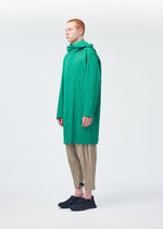 A person with short red hair wears HOMME PLISSÉ ISSEY MIYAKE’s PADDED PLEATS: a long, bright green textured coat with embroidered pattern, beige pleated pants, and black shoes, standing sideways against a plain white background.