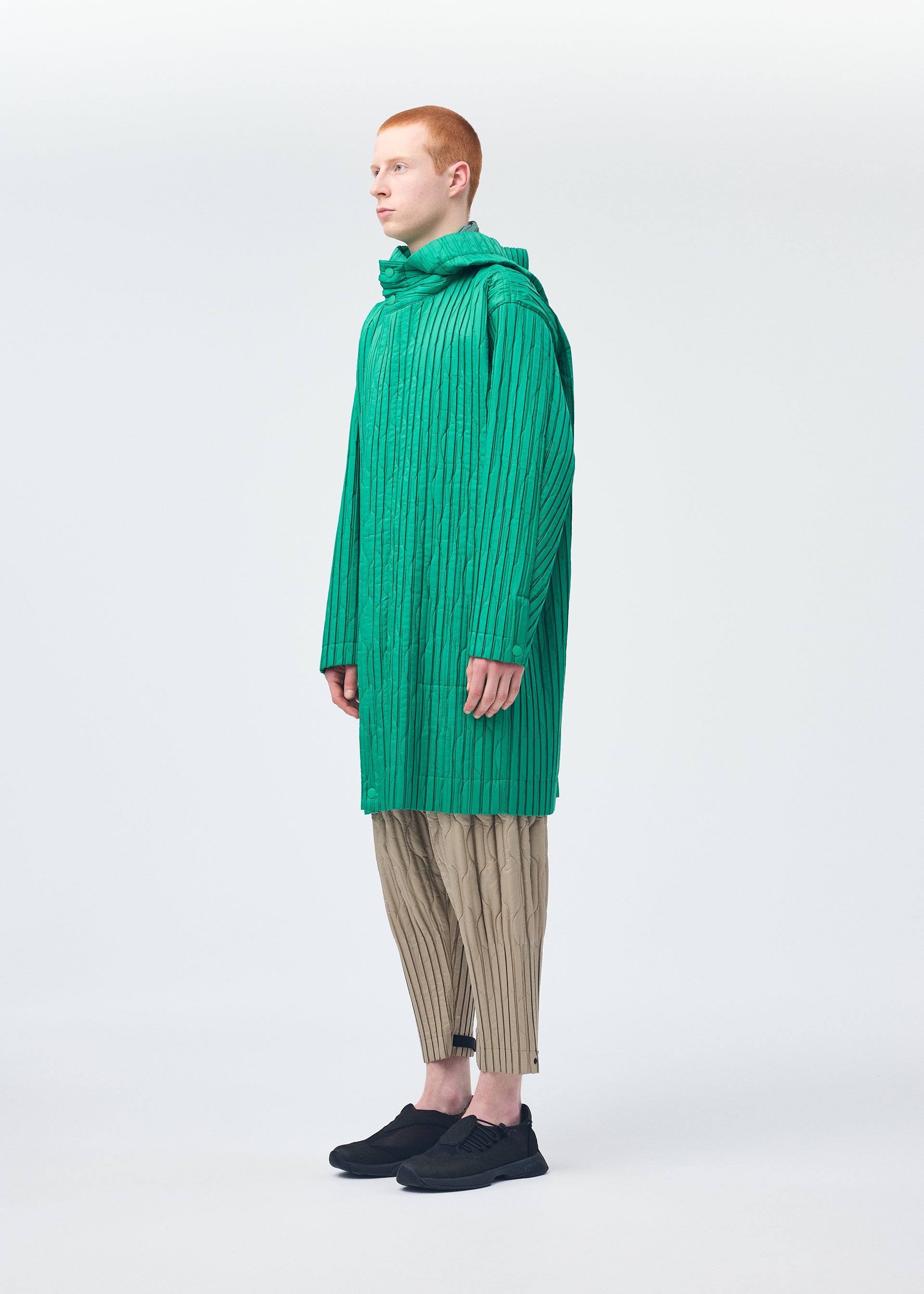 Zoomed view A person with short red hair wears HOMME PLISSÉ ISSEY MIYAKE’s PADDED PLEATS: a long, bright green textured coat with embroidered pattern, beige pleated pants, and black shoes, standing sideways against a plain white background.