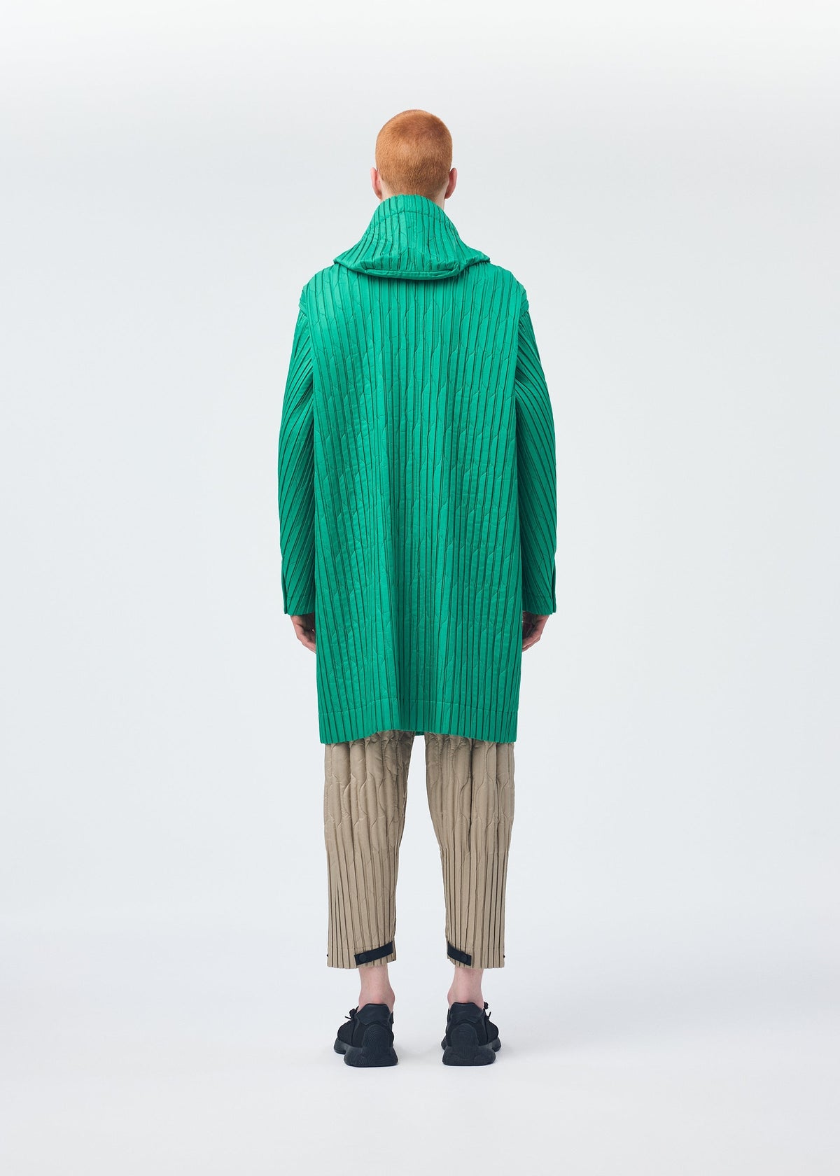 A person with short red hair, seen from behind, models the HOMME PLISSÉ ISSEY MIYAKE PADDED PLEATS: a bright green textured coat with embroidery, worn over beige pleated pants and black shoes against a white background. view 7 of 7