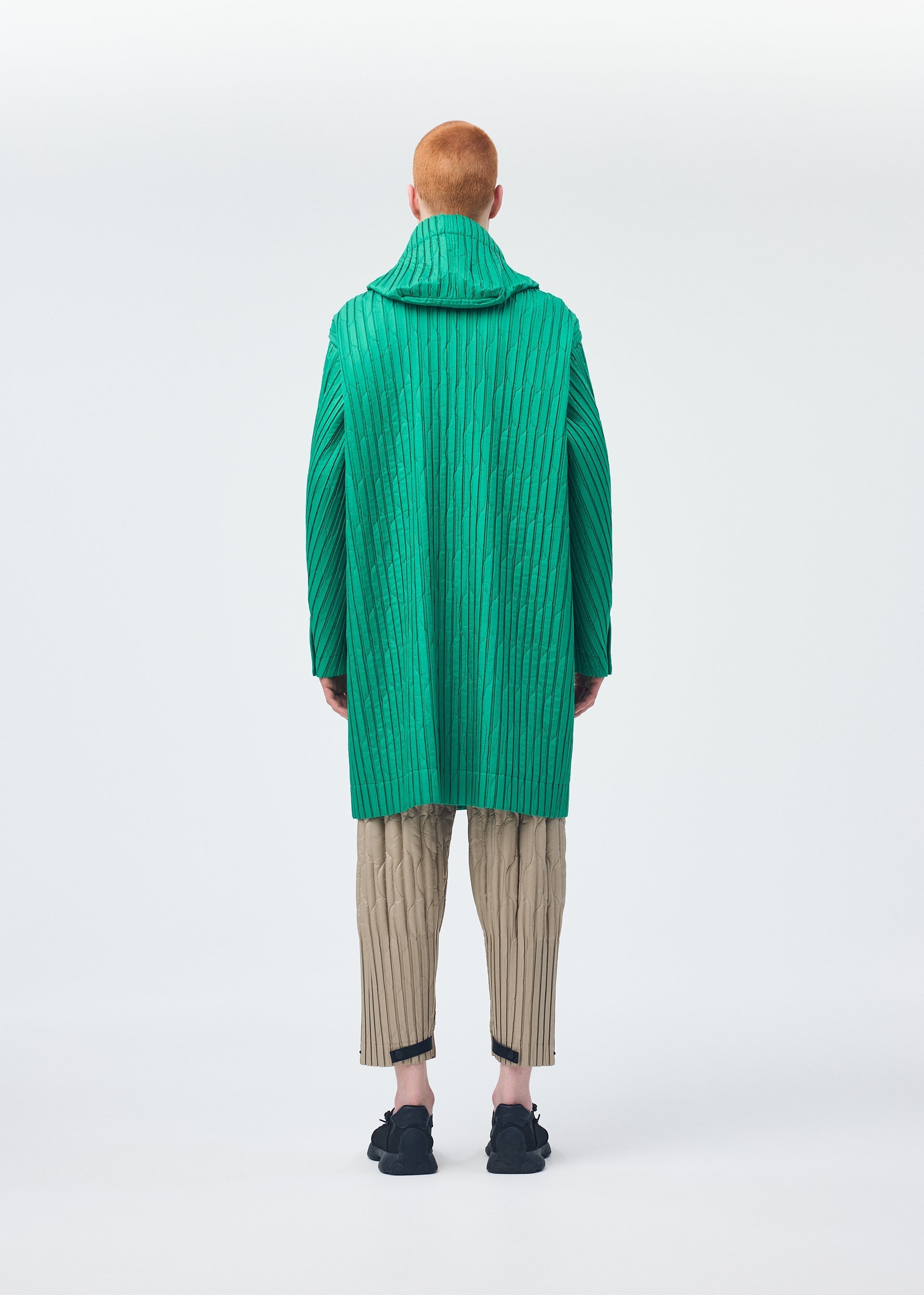 Zoomed view A person with short red hair, seen from behind, models the HOMME PLISSÉ ISSEY MIYAKE PADDED PLEATS: a bright green textured coat with embroidery, worn over beige pleated pants and black shoes against a white background.