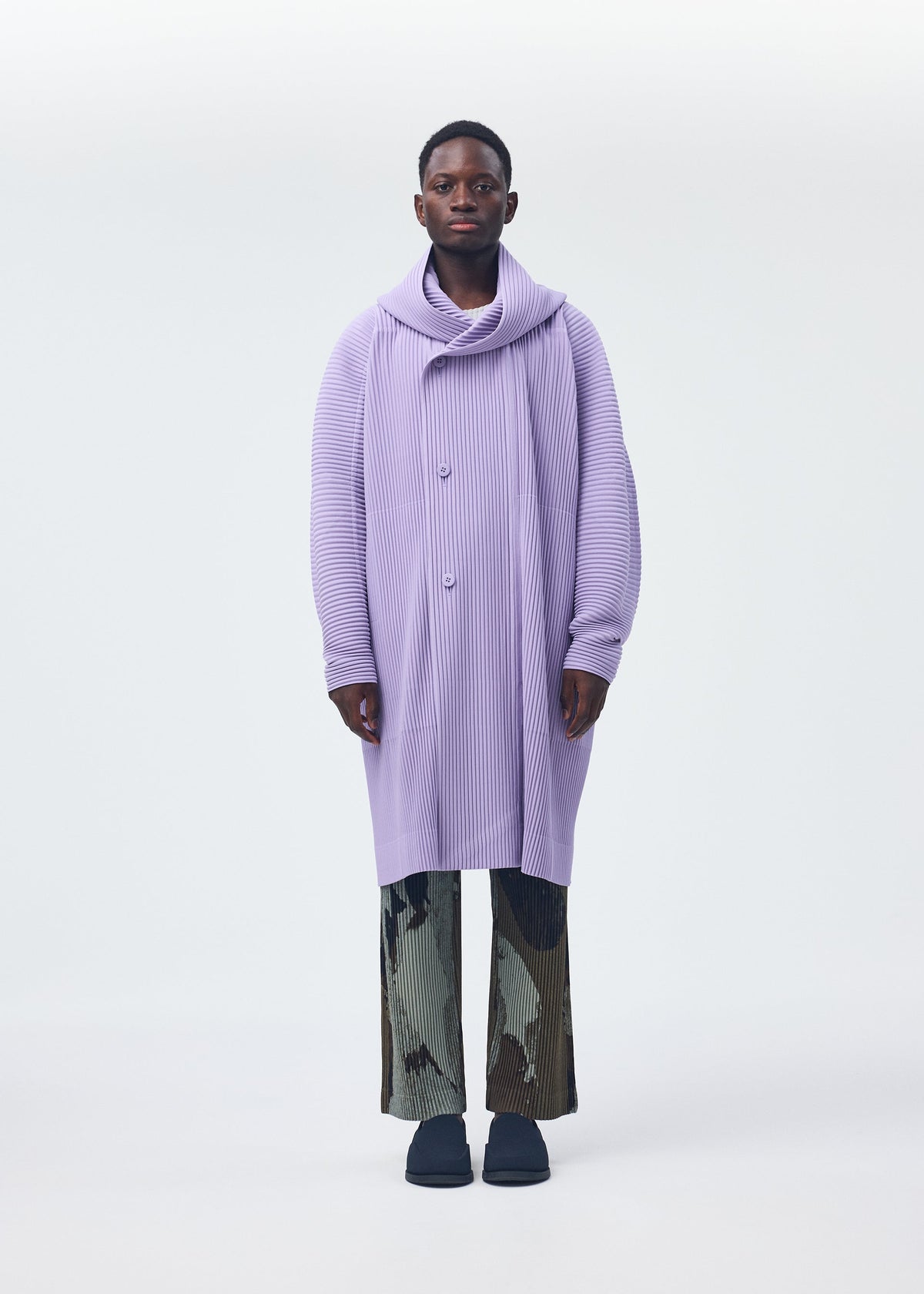 A person stands facing forward, wearing the HOMME PLISSÉ ISSEY MIYAKE MONTHLY COLORS : DECEMBER coat—long, oversized, ribbed lavender with a large cowl neck—paired with black-and-white patterned pants against a plain light background. view 4 of 6