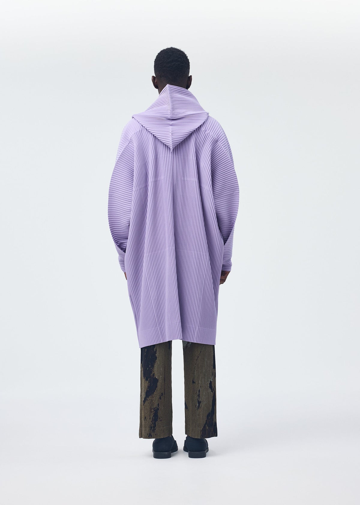 A person wears the HOMME PLISSÉ ISSEY MIYAKE MONTHLY COLORS : DECEMBER—a long, light purple hooded coat with textured vertical lines—over wide-legged, dark brown pants with a black abstract pattern, against a plain light background. view 6 of 6