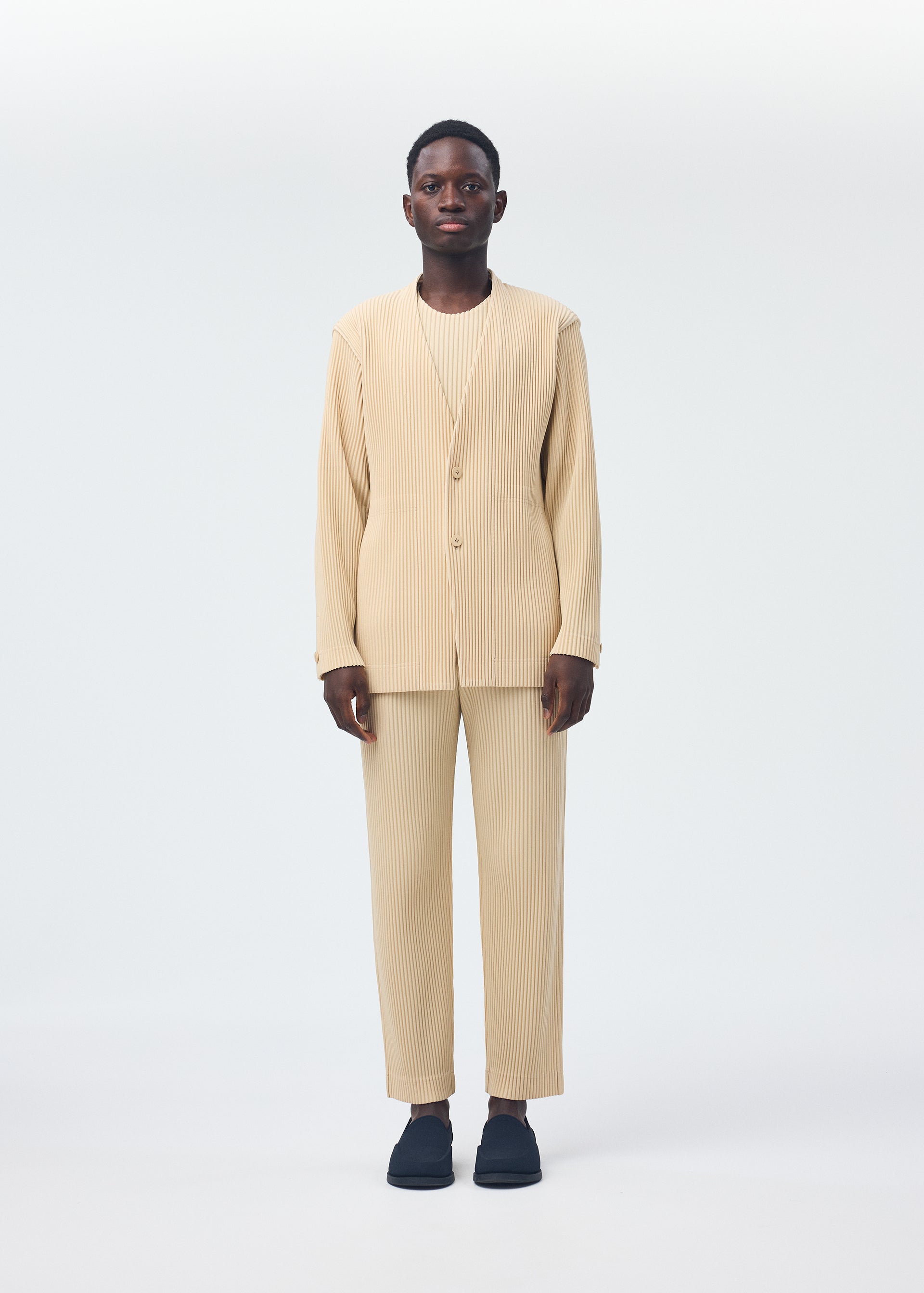 Zoomed view A person with short hair stands facing forward against a plain white background, wearing the beige TAILORED PLEATS 2 set from HOMME PLISSÉ ISSEY MIYAKE and black slip-on shoes. The outfit features a collarless pinstriped jacket, matching pants, and a shirt.