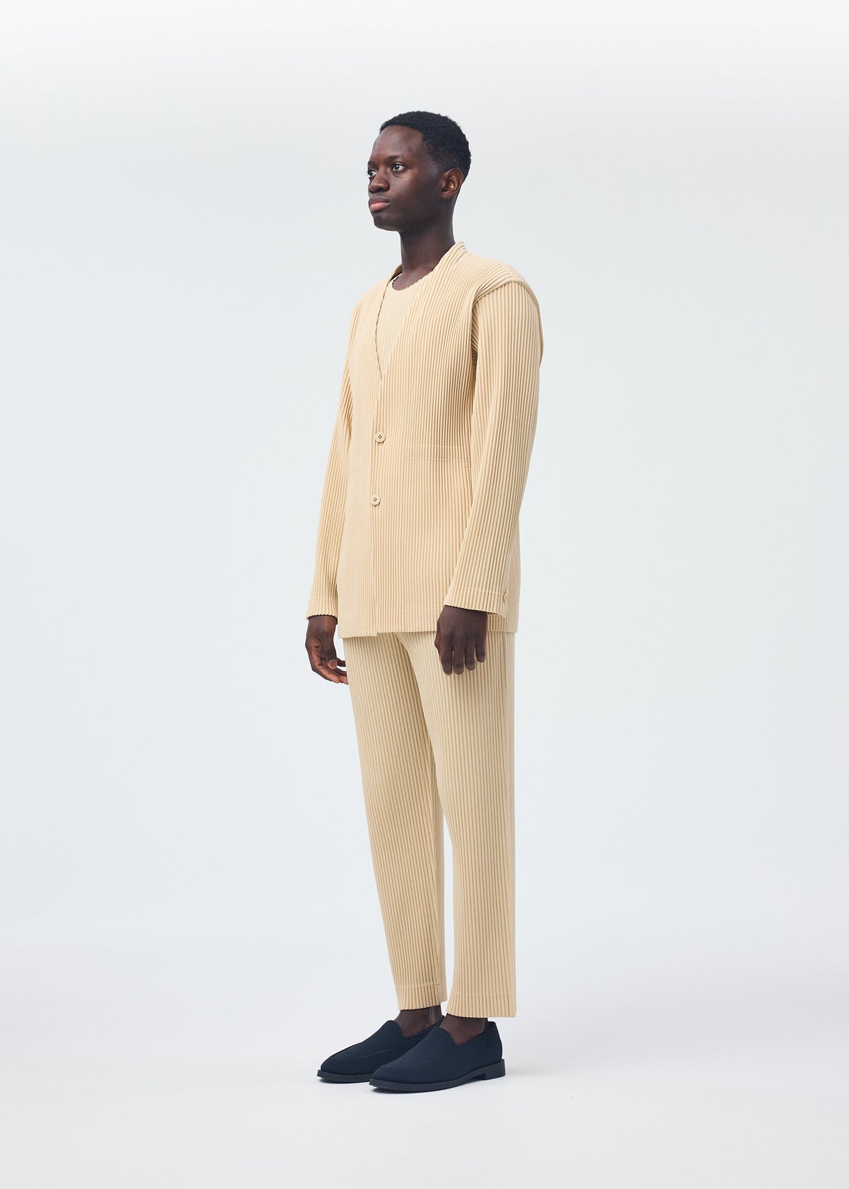 A person faces left with a neutral expression, wearing the HOMME PLISSÉ ISSEY MIYAKE TAILORED PLEATS 2 beige long-sleeved ribbed matching top and pants set in quick-drying polyester, paired with black slip-on shoes against a light background. view 6 of 7