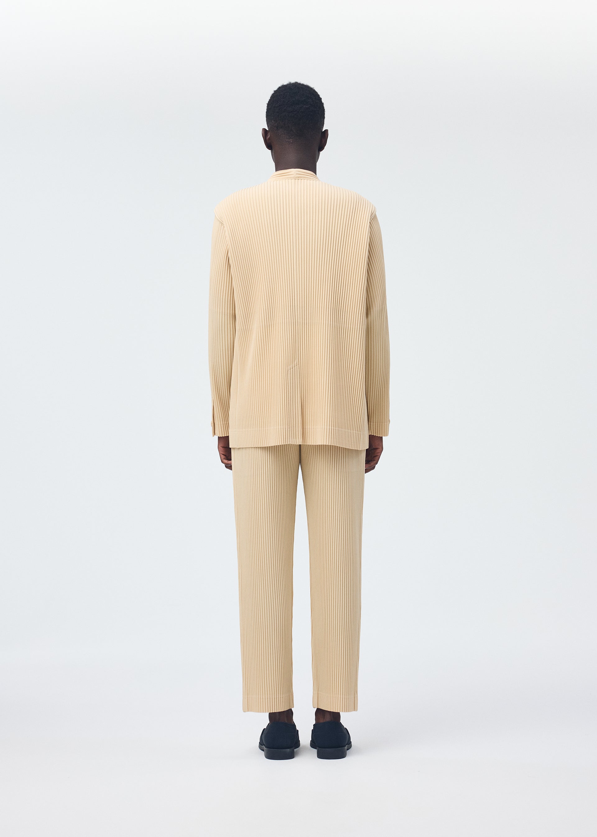 Zoomed view A person stands facing away, wearing the HOMME PLISSÉ ISSEY MIYAKE TAILORED PLEATS 2 beige pleated long-sleeve top and pants set, made from quick-drying polyester with black shoes, against a plain white background.
